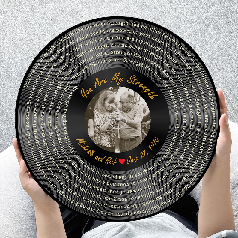 May include: A black circular vinyl record wall art piece featuring a central black and white photograph of a couple. The text "You Are My Strength" is printed in gold, with "Michelle and Rich" and "June 27, 1970" below the photo.