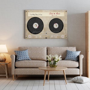 May include: A framed cassette tape design with the words "Can't Help Falling In Love" and names "Chris & Maria" with a date. The artwork hangs above a beige sofa with blue patterned pillows and a wooden coffee table.
