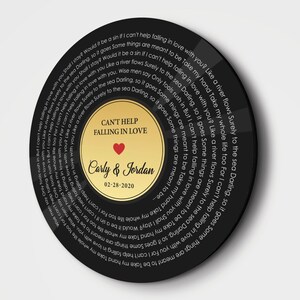 May include: A circular black wall art piece designed like a vinyl record. The center gold label features the text CAN'T HELP FALLING IN LOVE, a red heart, the names Carly & Jordan, and the date 02-28-2020. Lyrics spiral around the black surface.