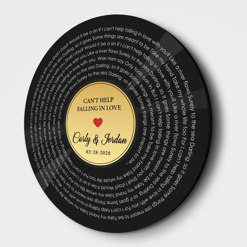 May include: A circular black wall art piece designed like a vinyl record. The center gold label features the text CAN'T HELP FALLING IN LOVE, a red heart, the names Carly & Jordan, and the date 02-28-2020. Lyrics spiral around the black surface.