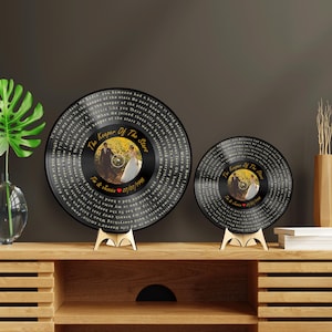 May include: Two circular black vinyl record wall art pieces on stands. Each features a central circular photograph of a couple in a field, the text "The Keeper Of The Stars," "Tim & Jessica," a red heart, "07/07/1995," and lyrics printed in white.