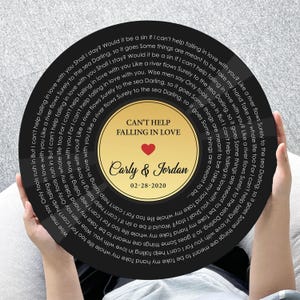 May include: A circular black wall art piece designed like a vinyl record featuring the lyrics to Can't Help Falling in Love printed in white. The center gold label displays the text Can't Help Falling in Love, Carly & Jordan, and 02-28-2020.