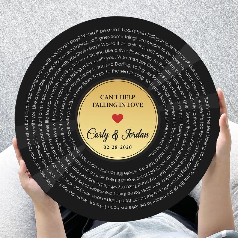 May include: A circular black wall art piece designed like a vinyl record featuring the lyrics to Can't Help Falling in Love printed in white. The center gold label displays the text Can't Help Falling in Love, Carly & Jordan, and 02-28-2020.