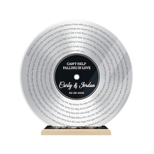 May include: A circular acrylic plaque designed like a vinyl record, featuring the text "CAN'T HELP FALLING IN LOVE", "Carly & Jordan", and the date "02-28-2020" in the center, with song lyrics printed in a spiral pattern on the clear outer edge.