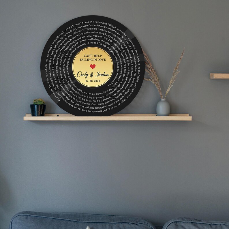 May include: A circular black wall plaque featuring the lyrics to Can't Help Falling in Love printed in a spiral pattern. The center is a gold circle with the text Can't Help Falling in Love, a red heart, the names Carly & Jordan, and 02-28-2020.