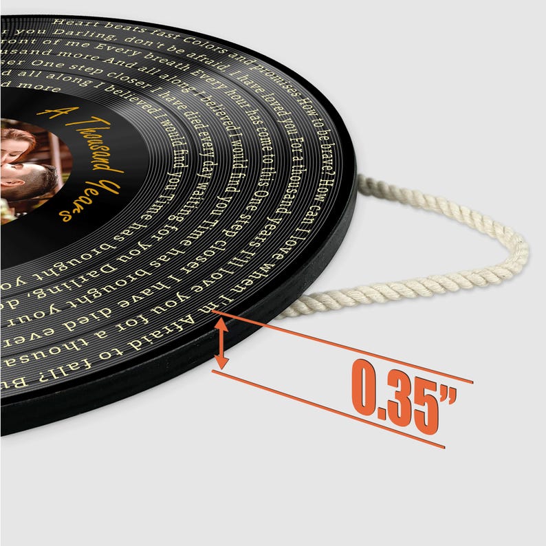 May include: A circular black wall plaque designed like a vinyl record, featuring a photograph of a couple and the text A Thousand Years. The lyrics are printed in white around the edge. A white rope is attached for hanging, and it is 0.35 inches thick.