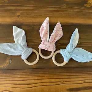 May include: Three wooden teethers with fabric bunny ears. One teether has blue and white polka dot ears, one has pink and white bunny ears with a pink and white floral pattern, and one has blue and white anchor print ears.
