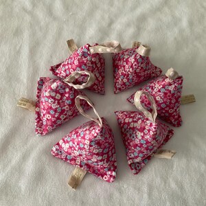 May include: Six pyramid-shaped fabric sachets featuring a vibrant pink floral pattern with small white and blue blossoms. Each sachet includes a small beige ribbon loop at the top and a tiny beige fabric tag attached to the side or bottom edge.