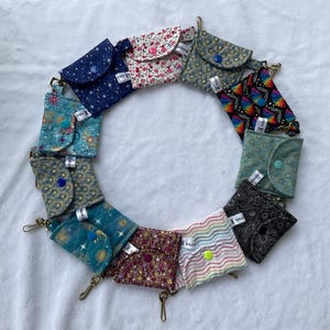May include: A circular arrangement of twelve small, square fabric pouches, each featuring a unique pattern including floral, celestial, geometric, and rainbow designs. Each pouch has a snap button closure and a small metal lobster clasp attached.