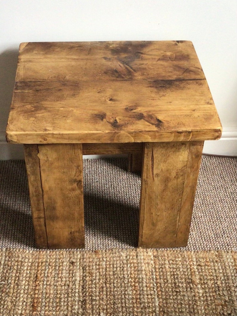 May include: A rustic wooden side table with a natural wood finish. The table has a square top and four legs. The table is sitting on a woven rug.