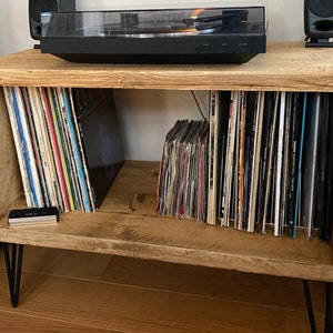 May include: A rustic wooden record player stand with black metal hairpin legs. The stand has two shelves for storing vinyl records. A black record player sits on top of the stand.