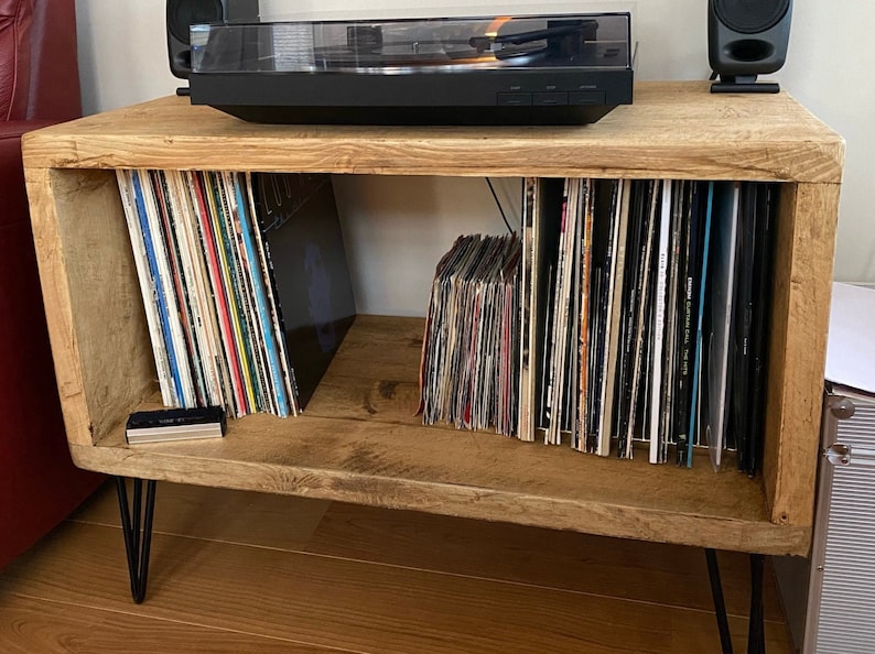 May include: A rustic wooden record player stand with black metal hairpin legs. The stand has two shelves for storing vinyl records. A black record player sits on top of the stand.