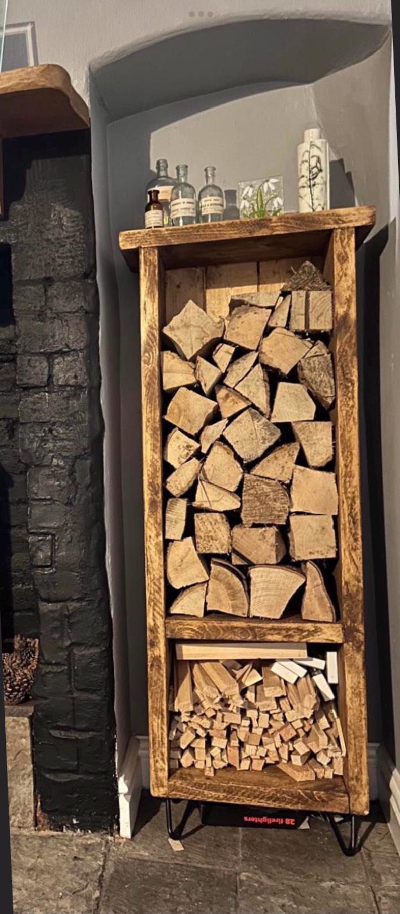 May include: A rustic wooden log rack with two shelves, one for larger logs and one for smaller kindling. The rack is stained a dark brown and has black hairpin legs. It is filled with stacked logs and kindling.