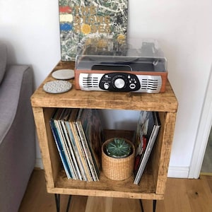 May include: A wooden record player stand with a record player on top and vinyl records in the shelves. The stand has hairpin legs and is painted a dark brown color.