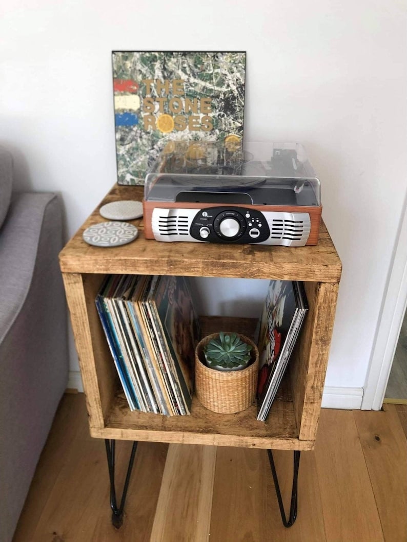 May include: A wooden record player stand with a record player on top and vinyl records in the shelves. The stand has hairpin legs and is painted a dark brown color.