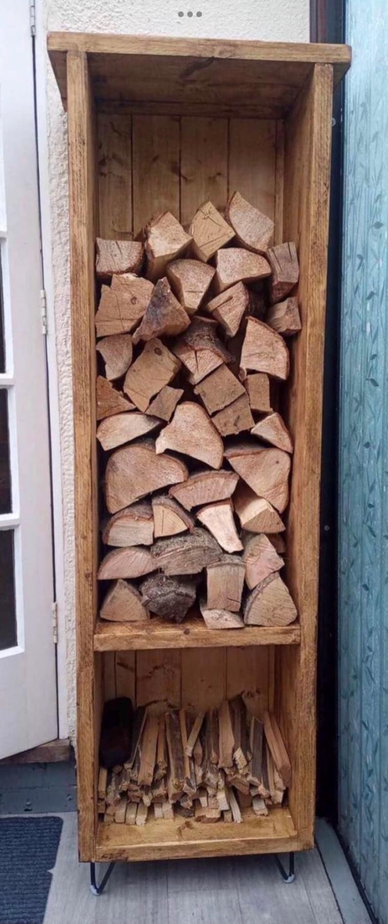 May include: A wooden log rack with two shelves. The top shelf is filled with stacked logs of wood. The bottom shelf is filled with smaller pieces of kindling. The rack is standing on four black metal hairpin legs.