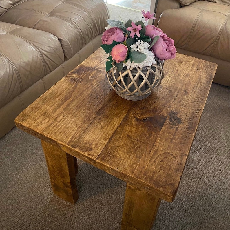 May include: A rustic wooden coffee table with a natural wood finish. The table has a square top and two thick legs. A wicker basket filled with pink and white flowers sits on the table.