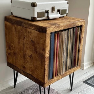 May include: A wooden record player stand with black metal hairpin legs. The stand has a shelf for a record player and a compartment for storing vinyl records. The stand is stained a dark brown color.