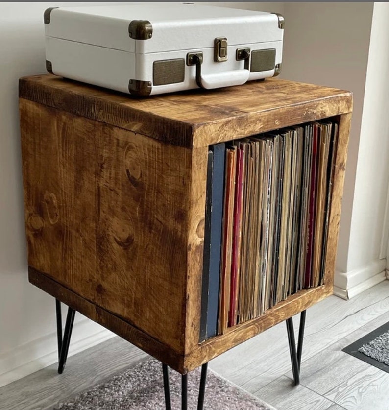 May include: A wooden record player stand with black metal hairpin legs. The stand has a shelf for a record player and a compartment for storing vinyl records. The stand is stained a dark brown color.
