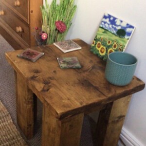 May include: A rustic wooden side table with a distressed brown finish. The table has four legs and a square top. There are four coasters on the table, two of which are visible in the image. A teal ceramic mug is on the table, and a framed print of a sunflower field is leaning against the wall behind the table.