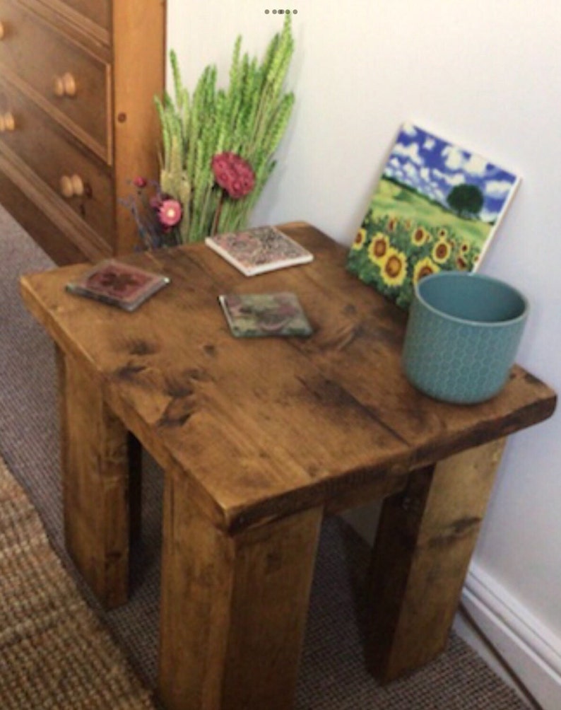 May include: A rustic wooden side table with a distressed brown finish. The table has four legs and a square top. There are four coasters on the table, two of which are visible in the image. A teal ceramic mug is on the table, and a framed print of a sunflower field is leaning against the wall behind the table.