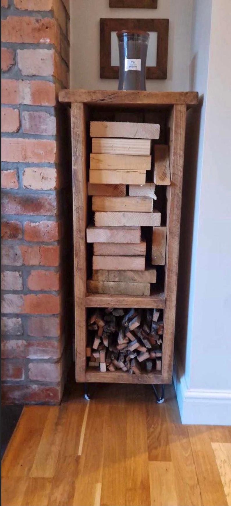May include: A rustic wooden log storage rack with two shelves. The top shelf is filled with stacked wooden planks, and the bottom shelf is filled with kindling. The rack is standing on four black metal legs.