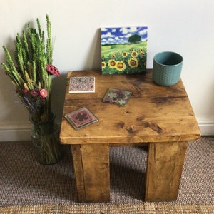 May include: A rustic wooden coffee table with a distressed finish. The table has four legs and a rectangular top. There are four coasters on the table, including one with a floral design and another with a sunflower design. A teal mug is on the table, and a vase with dried flowers is on the floor next to the table.
