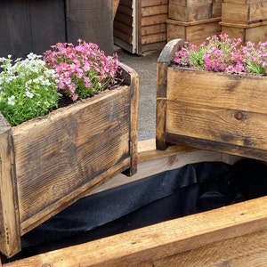 May include: Two wooden planter boxes with a black liner inside. The planter boxes are made of reclaimed wood and have a rustic look. One planter box is filled with pink flowers and the other is filled with white flowers.