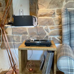 May include: A wooden side table with a record player on top and a stack of vinyl records on the shelf. The table has hairpin legs and a copper-colored lamp next to it.