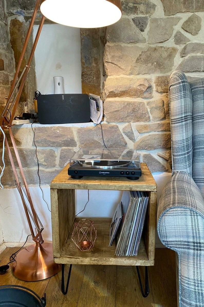 May include: A wooden side table with a record player on top and a stack of vinyl records on the shelf. The table has hairpin legs and a copper-colored lamp next to it.