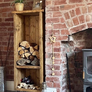 May include: A rustic wooden log rack with two shelves, black hairpin legs, and a small framed photo on top. The rack is filled with logs and is placed in front of a brick fireplace.