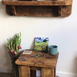 May include: A rustic wooden shelf with a decorative tile box and string lights. Below the shelf is a small wooden table with a sunflower tile, coasters, and a teal blue cup.