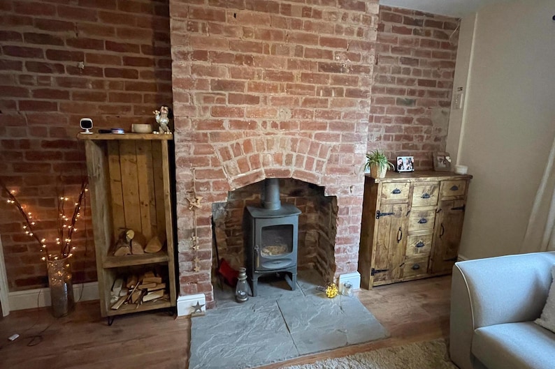 May include: A wood-burning fireplace with a black cast iron stove in a brick fireplace. The fireplace is in a living room with a wooden cabinet and a couch.