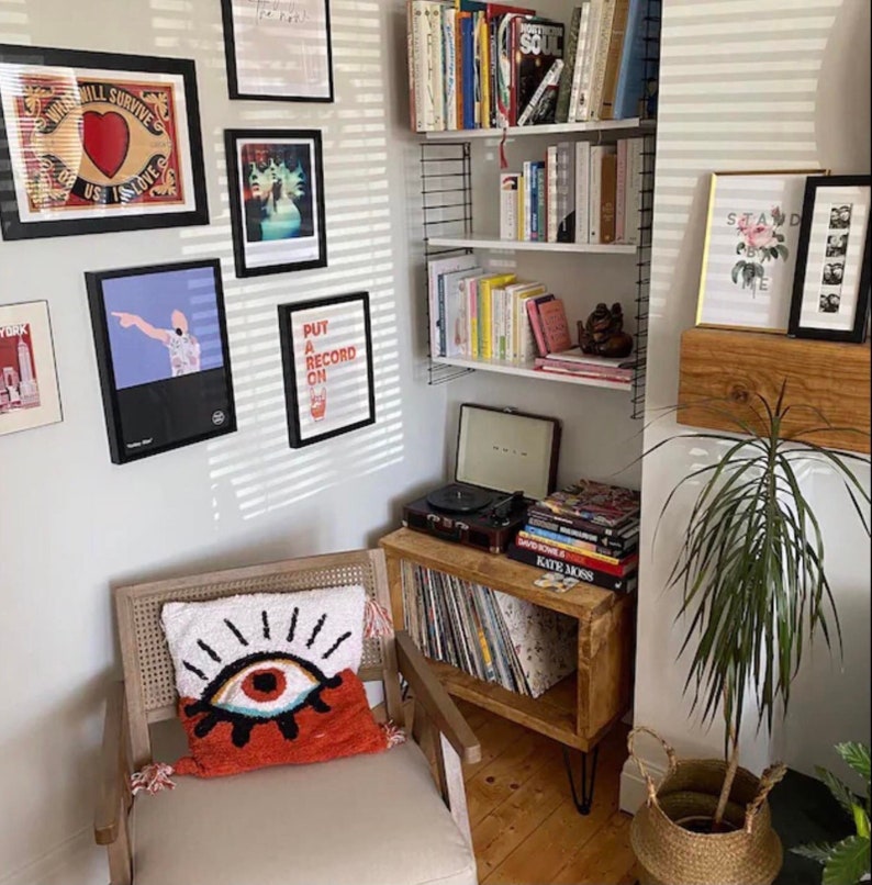 May include: A white armchair with a red and blue eye pillow. The chair is in a room with a record player, a bookshelf, and a plant. The bookshelf is filled with books and has a record player on the bottom shelf. The plant is in a wicker basket. The wall is white with a striped pattern. There are several framed pictures on the wall. One of the pictures says "Put a record on".