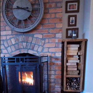 May include: A brick fireplace with a black metal fire screen and a burning fire. A large gray clock hangs on the wall above the fireplace. A wooden shelf unit with stacked firewood sits to the right of the fireplace.