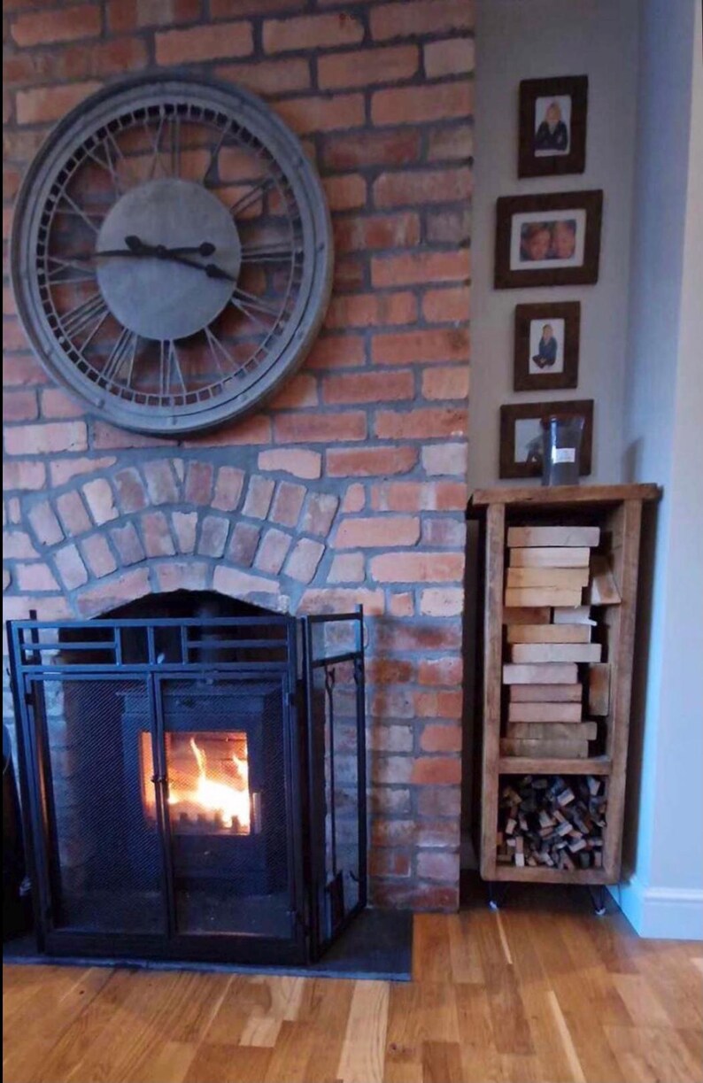 May include: A brick fireplace with a black metal fire screen and a burning fire. A large gray clock hangs on the wall above the fireplace. A wooden shelf unit with stacked firewood sits to the right of the fireplace.
