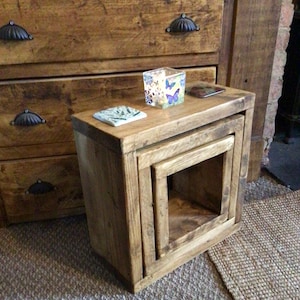 May include: A set of three nesting tables made from reclaimed wood. The tables are square and have a natural wood finish. The top table has a small candle holder and a decorative plate on it.