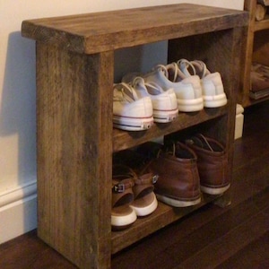 May include: A rustic wooden shoe rack with two shelves. The top shelf is a bench and the bottom shelf is for shoes. There are white trainers and brown boots on the shelves.