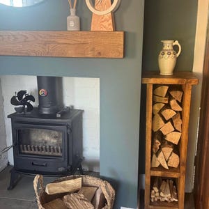 May include: A wood-burning fireplace with a black cast iron stove and a wood-burning stove fan. A wood-burning fireplace with a black cast iron stove and a wood-burning stove fan. There is a wooden shelf above the fireplace with a clock and a diffuser. A wooden log rack with a metal frame is next to the fireplace, filled with logs.