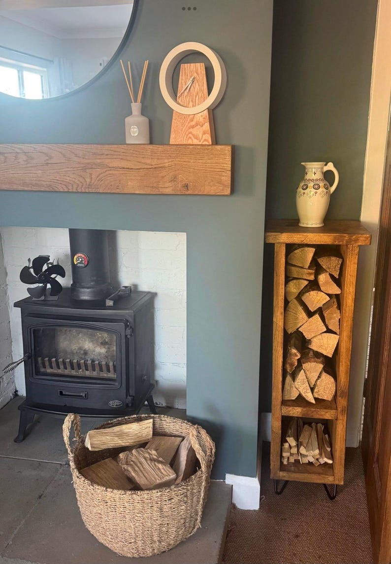 May include: A wood-burning fireplace with a black cast iron stove and a wood-burning stove fan. A wood-burning fireplace with a black cast iron stove and a wood-burning stove fan. There is a wooden shelf above the fireplace with a clock and a diffuser. A wooden log rack with a metal frame is next to the fireplace, filled with logs.