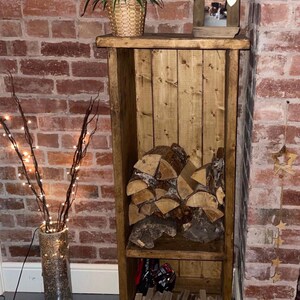 May include: A rustic wooden log storage rack with two shelves. The top shelf has a small framed photo and a potted plant. The bottom shelf has a stack of firewood and a few other items. The rack is standing on black hairpin legs.