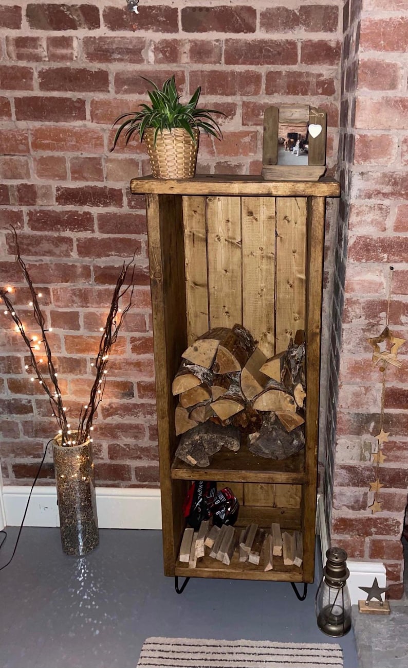 May include: A rustic wooden log storage rack with two shelves. The top shelf has a small framed photo and a potted plant. The bottom shelf has a stack of firewood and a few other items. The rack is standing on black hairpin legs.