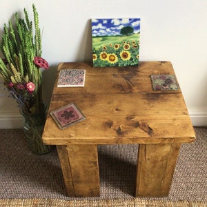 May include: A rustic wooden coffee table with a distressed finish. The table has four legs and a square top. There are three decorative coasters on the table, one with a floral design, one with a sunflower field design, and one with a geometric pattern.