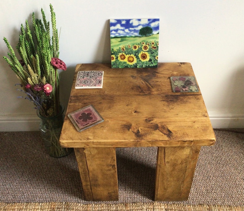 May include: A rustic wooden coffee table with a distressed finish. The table has four legs and a square top. There are three decorative coasters on the table, one with a floral design, one with a sunflower field design, and one with a geometric pattern.