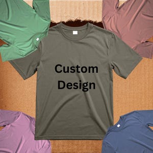 Könnte beinhalten: Ein olivgrünes T-Shirt mit den schwarzen Worten "Custom Design". Das Shirt ist von anderen T-Shirts in verschiedenen Farben umgeben, darunter Grün, Rosa und Blau. Die Shirts befinden sich auf einem braunen Hintergrund.