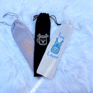 Puede incluir: Tres bolsas de cordón para llevar pajitas reutilizables. Las bolsas están hechas de diferentes telas y tienen diferentes diseños. Una bolsa es gris con una taza de té boba de dibujos animados, otra es negra con una taza de dibujos animados con una cara, y otra es beige con el texto "Tea Lover" y un tarro de albañil de dibujos animados.