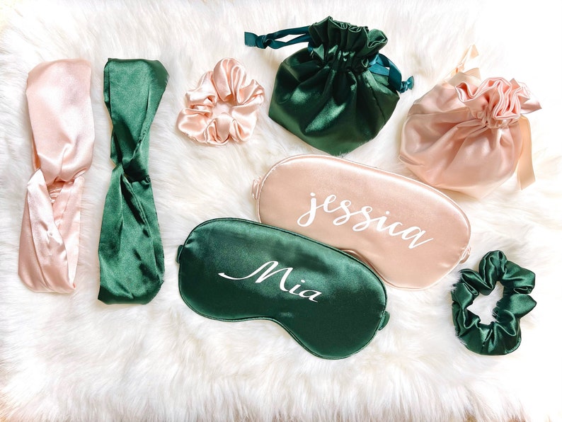 Op de afbeelding: Een assortiment satijnen slaapaccessoires in roze en smaragdgroen. De items omvatten haarbanden, scrunchies, zakjes met trekkoord en gepersonaliseerde slaapmaskers met namen als "Jessica" en "Mia" in witte letters.