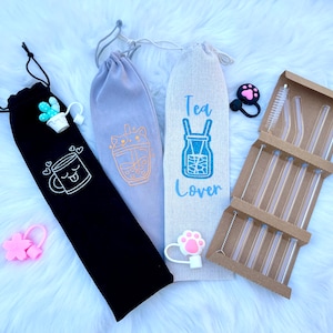 Puede incluir: Tres bolsas de tela en negro, gris y beige con diferentes diseños bordados. Las bolsas son para guardar pajitas de beber de vidrio reutilizables. Las pajitas están en un soporte de cartón marrón con un cepillo de limpieza. Las bolsas tienen diseños de dibujos animados lindos, que incluyen una taza de té, un té de boba y un tarro de té. El texto en las bolsas dice "Tea Lover".