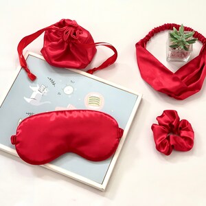 Op de afbeelding: Een rode satijnen slaapset met een oogmasker, scrunchie, hoofdband en trekkoordtas. De set wordt gepresenteerd op een wit oppervlak met een ingelijste illustratie op de achtergrond. De hoofdband heeft een kruisontwerp.