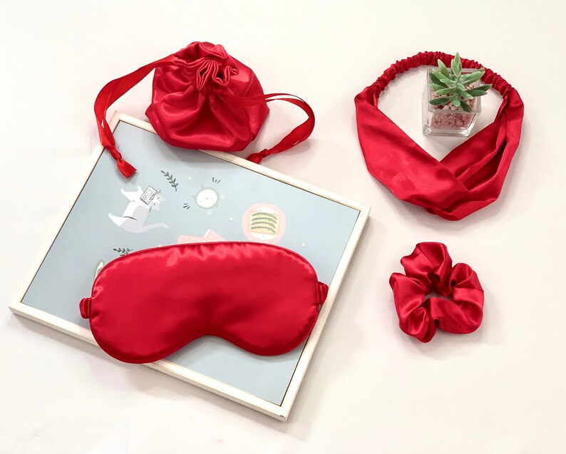 Op de afbeelding: Een rode satijnen slaapset met een oogmasker, scrunchie, hoofdband en trekkoordtas. De set wordt gepresenteerd op een wit oppervlak met een ingelijste illustratie op de achtergrond. De hoofdband heeft een kruisontwerp.